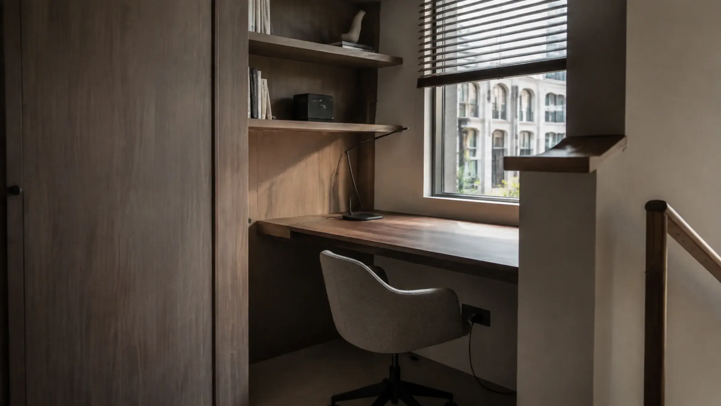 Compact home office built into an alcove with a narrow floating desk and a single task lamp, daylight through a partly open blind