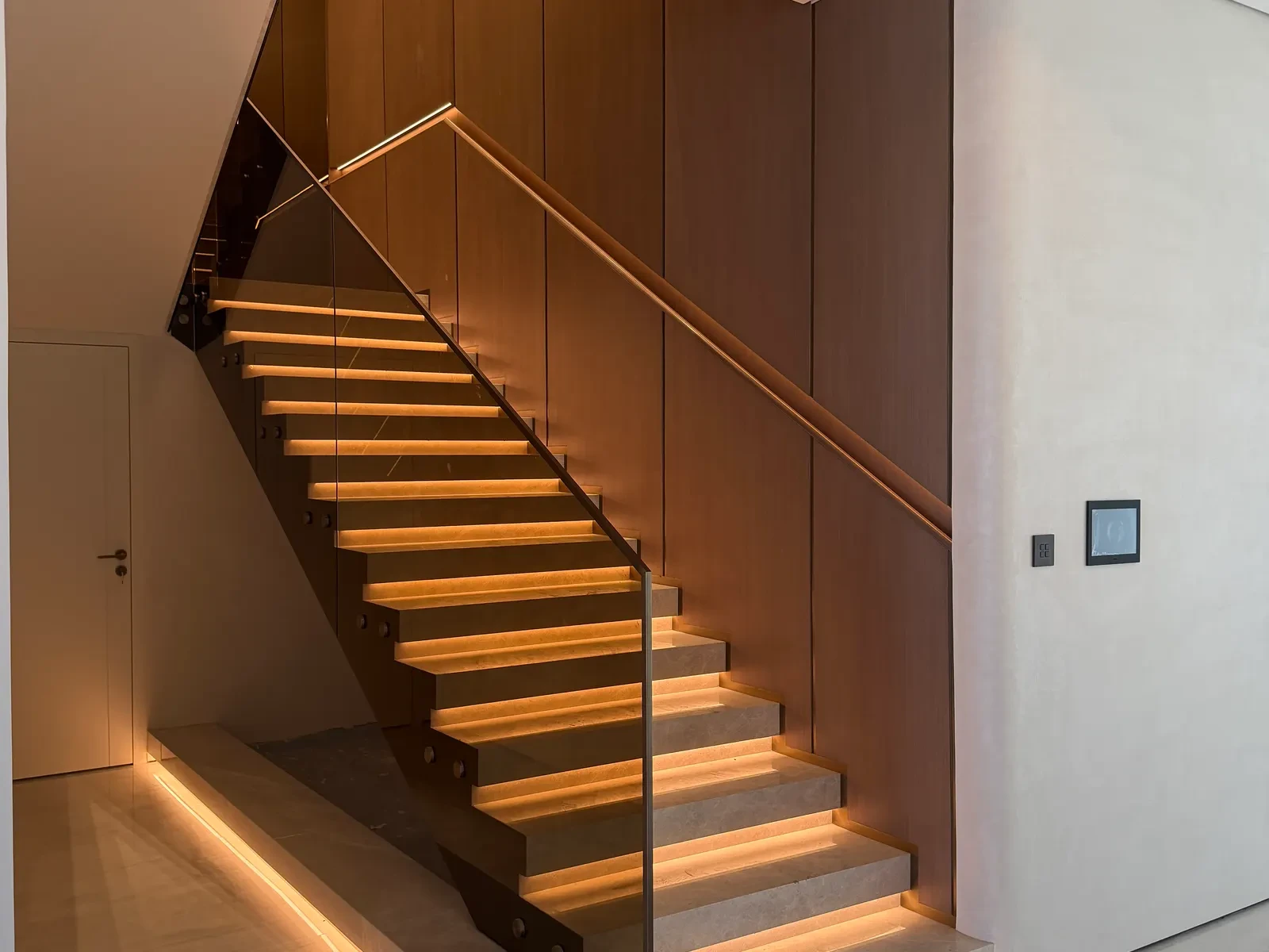 Floating timber stair with lit treads and a glass balustrade against timber panelling